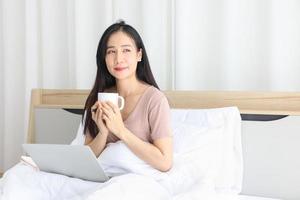 mujer asiática que trabaja en línea desde casa con una computadora portátil mientras toma café en su cama para una nueva vida normal durante el período de cuarentena de covid-19 foto