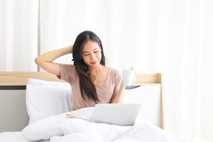 Young pretty Asian girl working from home on her bed using computer laptop to access her work online with copy space photo