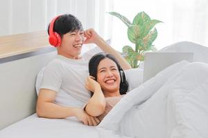 Young Asian couple lying in bed watching and laughing over comedy movie together in bedroom photo