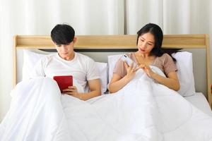 Asian couple looking at each other smartphone for social media while laying down in the bed planning for their upcoming wedding photo