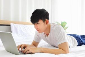 Young freelance Asian man working from home using laptop computer while sitting on the bed with copy space photo