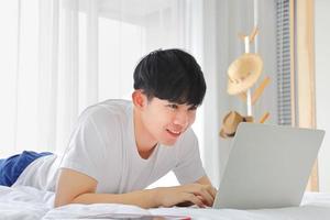 Young freelance Asian man working from home using laptop computer while sitting on the bed photo