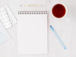 open notebook with blank white clean page to write the treatment plan of the illnedd. Pills, pen, paper, thermometer on white wood background, top view. photo