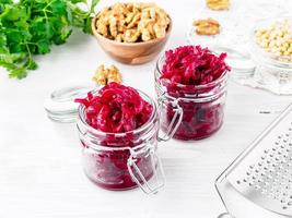 Fresh salad of grated boiled beetroot in jars, white wooden background, side view.