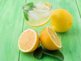 limonada en un vaso, medio limón, hojas frescas sobre la mesa verde. una refrescante bebida fría de agua con hielo, menta y rodajas de limón foto