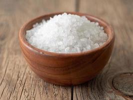 White bowl with large sea salt on brown bright wooden table. Top view, close up, selective focus photo