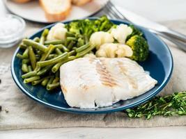 Boiled sea fish cod fillet with vegetables on blue plate, grey wooden table, side view. Healthy diet photo