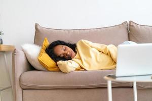 mujer afroamericana aburrida durmiendo en el sofá en la sala de estar mientras se reproduce una película en la computadora portátil foto