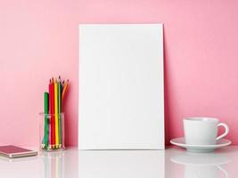 Blank white frame and crayon in jar, cup of coffee r tea on a white table against the pink wall with copy space photo