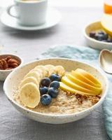 Whole oatmeal, large bowl of porridge with banana, blueberries, apple, nuts. Side view, vertical photo