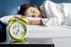 Beautiful young woman sleeping and smiling while lying in bed comfortably and blissfully on the background of alarm clock is going to ring. Relaxation and people lifestyles of tired worker. photo