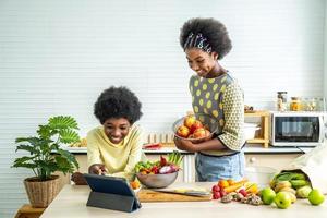 dos niños afroamericanos disfrutan cocinando juntos en una cocina moderna, dos hermanos felices hacen ensaladas de frutas y verduras a partir de un video tutorial de una tableta. aprende a cocinar en casa por videollamada foto