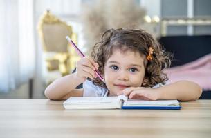 linda niña sentada en el escritorio en casa haciendo deberes, leyendo, escribiendo y pintando. los niños pintan. los niños dibujan niño en edad preescolar con libros en casa. los preescolares aprenden a escribir y leer. niño creativo foto