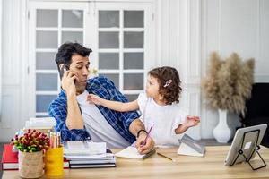 padre soltero de negocios tratando de trabajar haciendo una llamada telefónica mientras cuida a su hija juntos en casa, mientras que a la pequeña le gusta meterse con su padre mientras trabaja foto