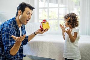 Happy father presenting gift box to excited cute daughterand cute daughter, Young father greeting his smiling adorable  girl, hugging him and giving gift box, bedroom interior photo