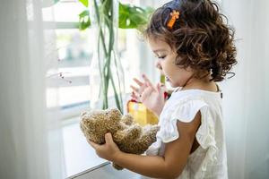 Happy little child girl stands by the window and hugs a favorite teddy bear photo