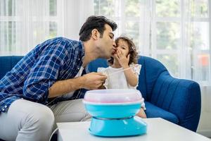 Caucasian family young father kissing his little daughter at mouth after eat cotton candy together on sofa at new house