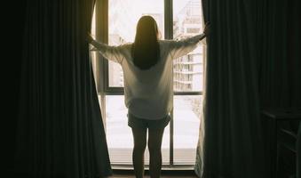 una joven y bella mujer encantada de pie junto al dormitorio y abriendo las cortinas, mira a la distancia de la ventana y recibe la bienvenida al nuevo día. las mujeres sonrientes se sienten emocionadas por las oportunidades o perspectivas de carrera en la vida. foto