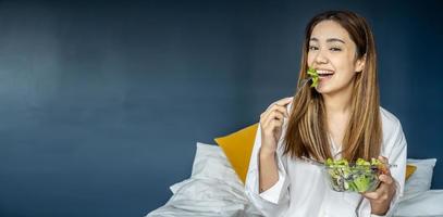 Gorgeous young woman sitting on bed eating vegetable salad, Balanced raw food diet. Positive girl having vegan lunch, taking care of her wellbeing