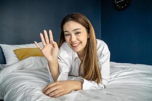 retrato de una chica alegre y atractiva que usa pijamas sonriendo alegremente y mira la cámara en un dormitorio acogedor en casa felicidad y alegría, saludando con la mano, dándole la bienvenida y saludándolo. foto