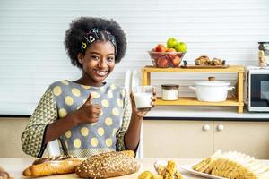 joven mujer feliz afroamericana pelo afro bebiendo un vaso de leche en casa feliz con una gran sonrisa haciendo bien el signo, pulgar hacia arriba con los dedos, excelente signo foto