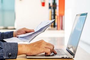 hombre de negocios revisando un informe o documento escrito mientras se sienta a trabajar en papeleo en una computadora portátil, pagando facturas en línea en una computadora portátil