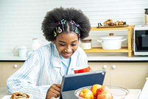 mujer joven afroamericana en la cocina mientras desayuna y toma té o leche en la taza envía un mensaje o llama con la tableta y sonrisas, concepto de red social, mensaje, tecnología foto