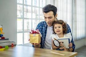 Happy father presenting gift box to excited cute daughterand cute daughter, Young father greeting his smiling adorable  girl, hugging him and giving gift box, bedroom interior photo