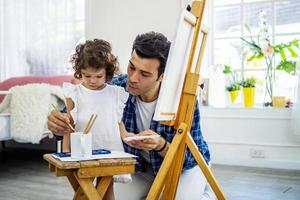 Happy family of father and kid drawing picture, using different paints and brushes. Handsome father and pretty little daughter against oil picture smiling, having fun. Lesson of art