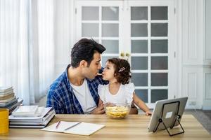 padre joven de familia caucásica comiendo papas fritas en la boca con una hija pequeña juntos y viendo dibujos animados programa educativo en línea foto