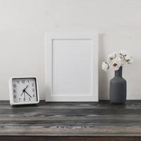 White frame, flower in vaze, clock on dark gray wooden table against the white wall with copy space. Mock up.