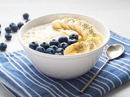 avena con plátanos, arándanos, chía, mermelada, miel, servilleta azul sobre fondo blanco de madera, vista lateral foto