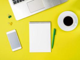 Top view of modern bright yellow office desktop with blank notepad, computer, smartphone. Mock up, empty space photo