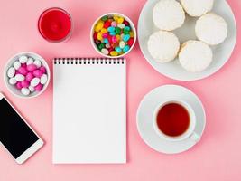 Cup of tea, notepad, mobile and plates of different sweets - marshmallows, lollipops, candy. Pink background, top view, flat lay. photo