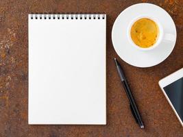 sábana blanca limpia en una libreta abierta en espiral, bolígrafo, teléfono móvil y taza de café en la plancha de la mesa de metal oxidado, vista superior foto