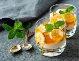 Healthy layered dessert with yogurt, banana, mango jam, cookie in glass on gray stone background, side view photo