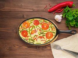 frying pan with Frittata in the center on a brown wooden table, forks, parsley, pepper, tomatoes, top view photo