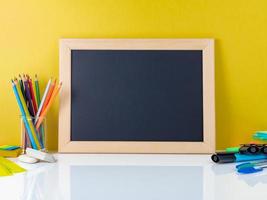 Chalkboard and school supplies on white table by the yellow wall. Side view, empty space. Back to school concept. photo