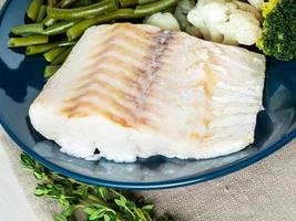 Baked sea fish cod fillet with vegetables on blue plate, gray napkin, side view, close up. Healthy diet photo