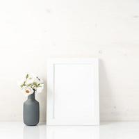 Blank white frame, flower in vaze on a white table against white wall with copy space. Mock up.