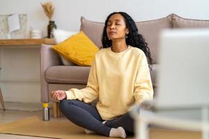Young african-american female sitting in lotus pose on mat in living room while meditating