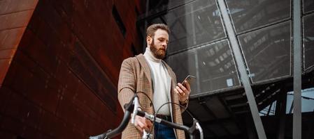 joven con bicicleta sosteniendo un teléfono inteligente frente al edificio. hombre de negocios creativo en un área de negocios moderna.