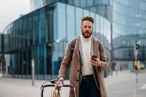 Portrait of hipster businessman with bike, using mobile and looking at camera. Business centre location. Coming to work