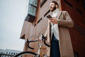 joven con bicicleta sosteniendo un teléfono inteligente frente al edificio. hombre de negocios creativo en un área de negocios moderna.