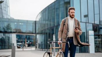 Portrait of hipster businessman with bike, using smartphone. Business centre location. Going from work. photo