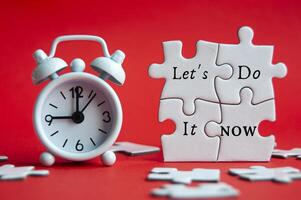 Let's do it now text on jigsaw puzzle with alarm clock pointing at 9 am on red cover background. photo