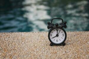 despertador negro sobre suelo de mármol con fondo de piscina borroso. el reloj puesto a las 8 en punto. foto