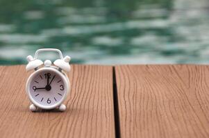 Alarm clock on wooden floor with blurred swimming pool background. The clock set at 9 o'clock. Morning concept. photo
