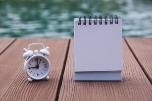 Alarm clock on wooden floor with empty notepad. With swimming pool background. The clock set at 9 o'clock. photo