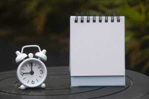 Alarm clock pointing at 9 o'clock with empty notepad. Blurred park background. Copy space concept